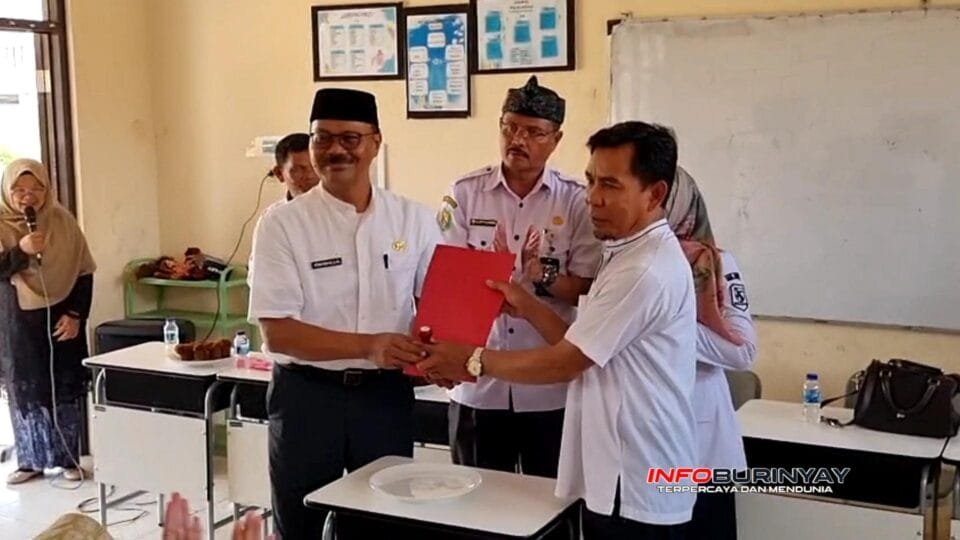Seorang pejabat sekolah menyerahkan dokumen kepada penerima dalam acara serah terima jabatan di ruang kelas, disaksikan para guru